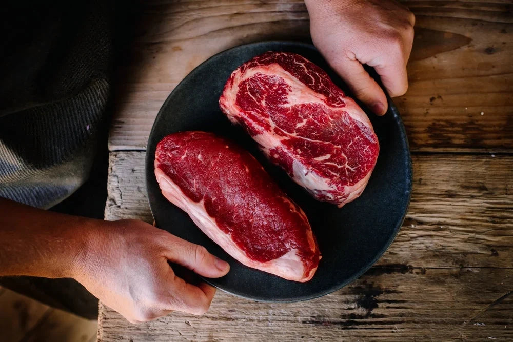 A black plate with a large sirloin steak and large ribeye steak - both raw.