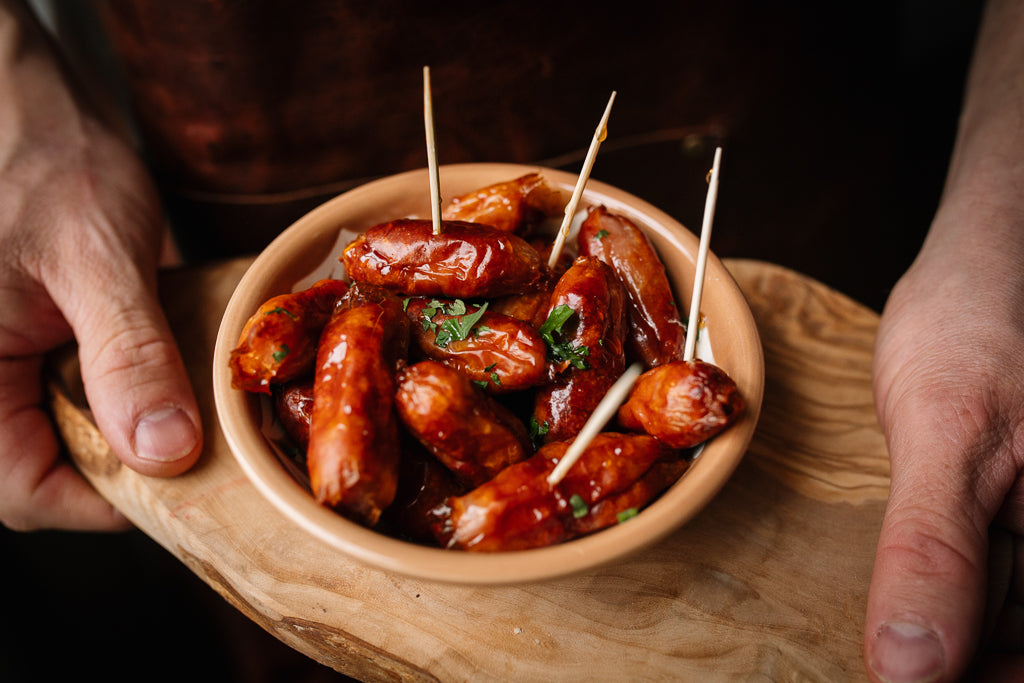 A close-up of a wooden bowl filled with glossy glazed sausage bites on a wooden board, held by two hands against a dark background.