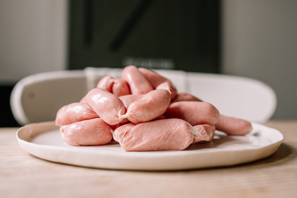 A pile of raw pink sausages tied in links, presented on a round white ceramic plate on a light wooden table.