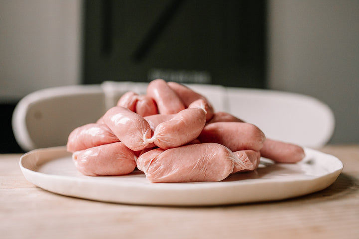 A pile of raw pink sausages tied in links, presented on a round white ceramic plate on a light wooden table.