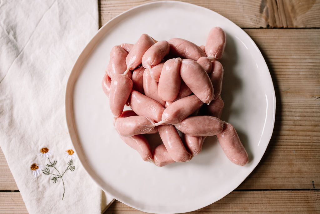 Raw pink sausages tied in a bundle, presented on a white ceramic plate over a rustic wooden table with a white embroidered cloth.