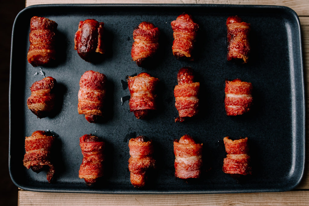 Twelve bacon-wrapped sausage rolls arranged in three rows on a dark rectangular baking sheet, photographed from above on a wooden surface.