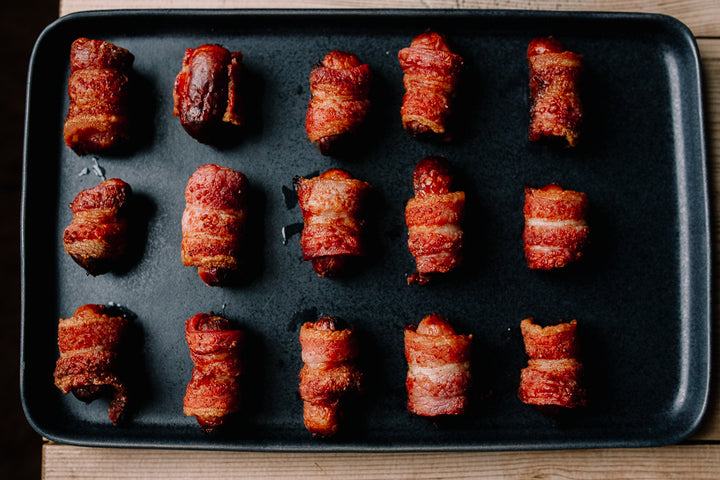 Twelve bacon-wrapped sausage rolls arranged in three rows on a dark rectangular baking sheet, photographed from above on a wooden surface.