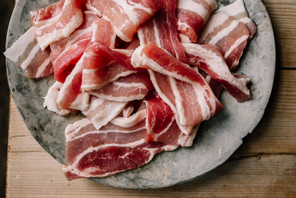 Raw sliced streaky bacon with white fat layers, piled on a round gray stone plate over a wooden surface.