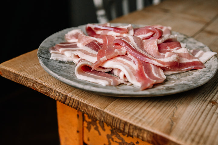Slices of raw bacon piled on a round gray plate, set on a rustic wooden table.