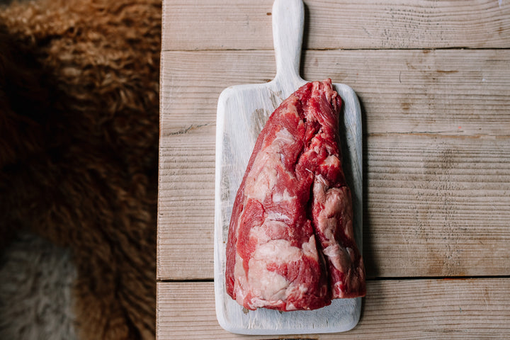 A raw cut of beef fillet tail, placed on a white rectangular cutting board with a handle, set on a light wood plank surface.