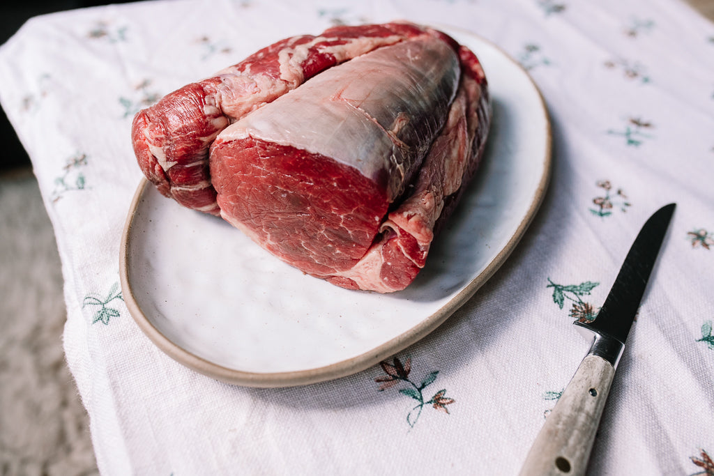 Raw beef chateaubriand roast on a white ceramic plate with a knife on a white floral tablecloth.