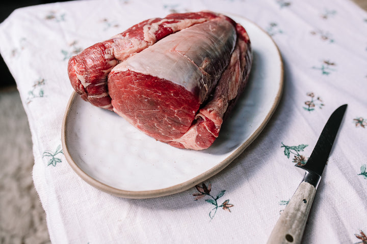 Raw beef chateaubriand roast on a white ceramic plate with a knife on a white floral tablecloth.