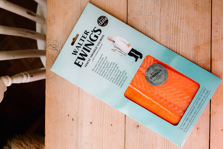 A package of Walter Ewing's Finely Smoked Salmon on a light wood tabletop. The packaging is light blue with a clear window showing the orange salmon inside, and the brand name 'Walter Ewing's' is clearly printed at the top.