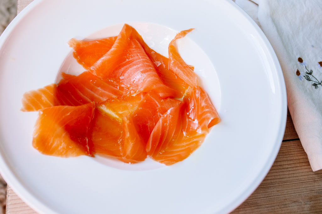 Thin slices of orange-pink salmon served on a white plate, set on a wooden surface with a folded white napkin partially visible at the right edge.