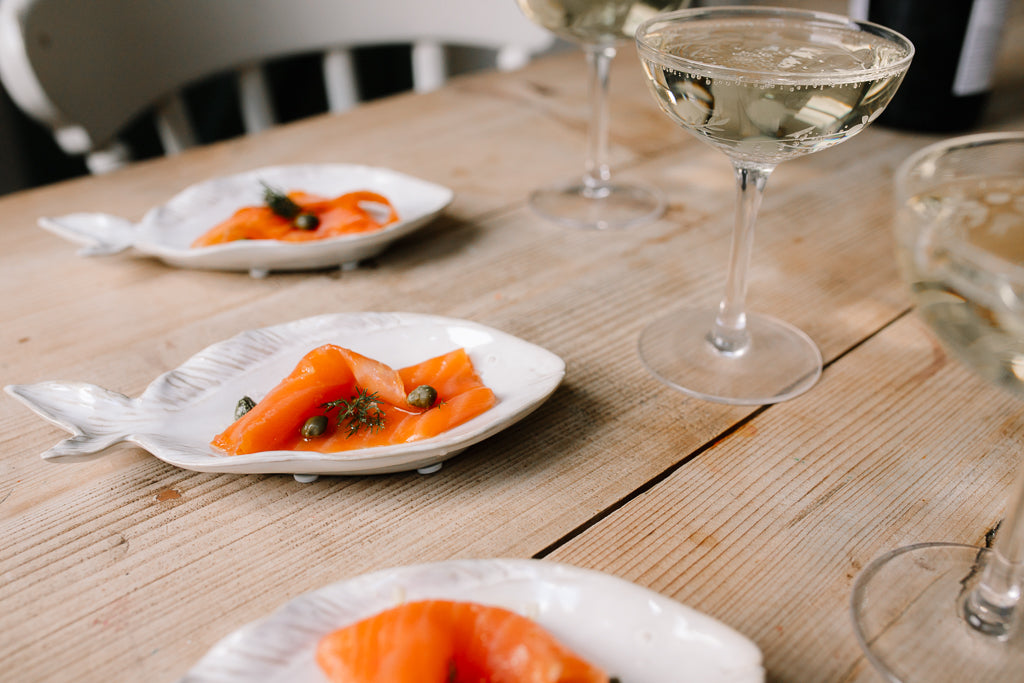 White fish-shaped plates with scalloped edges, each holding slices of orange salmon garnished with capers, set on a rustic wooden table alongside wine glasses.