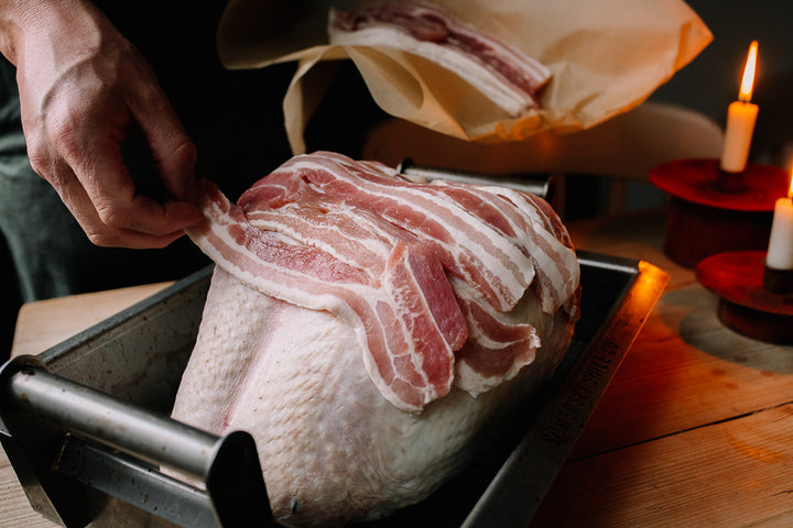 Man laying raw bacon on a raw turkey. Candles in the background. 