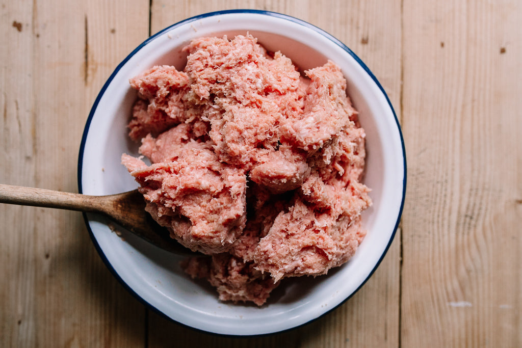 Raw ground pork mince for sausages or stuffing in a white enamel bowl with a blue rim, resting on a light wood plank surface with a wooden spoon visible.