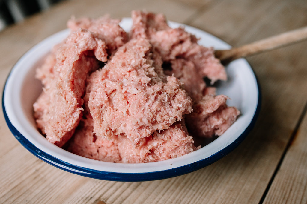 Raw ground Idevour Pork Sausage Mince in a white enamel bowl with a blue rim, sitting on a wooden table with a wooden spoon.