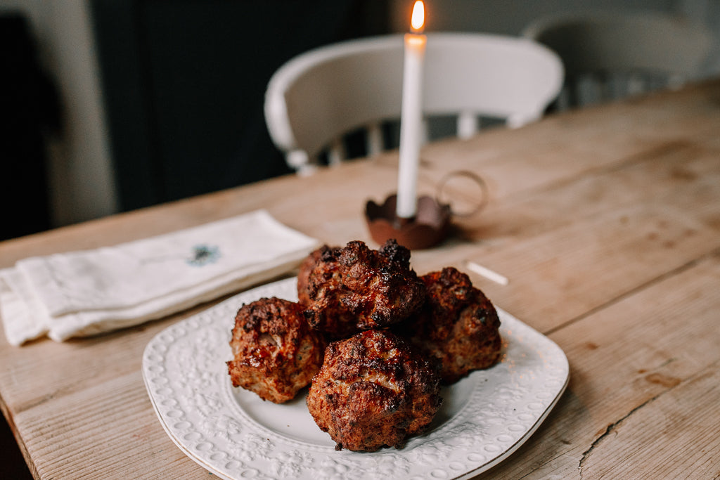 Five golden-brown baked pork stuffing balls on a white textured ceramic plate, set on a rustic wooden table with a lit white candle in the background.