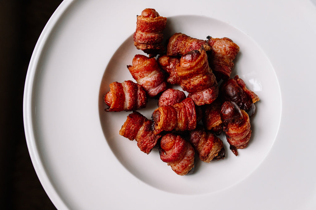 Crispy bacon-wrapped cocktail sausages (pigs in blankets) served on a white plate against a dark background.