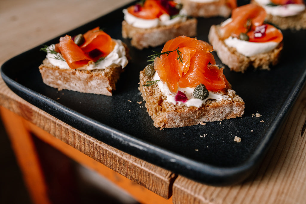 Six gourmet canapes on a black rectangular serving tray, featuring smoked salmon, cream cheese, capers, and dill on rustic bread, set on a wooden table.