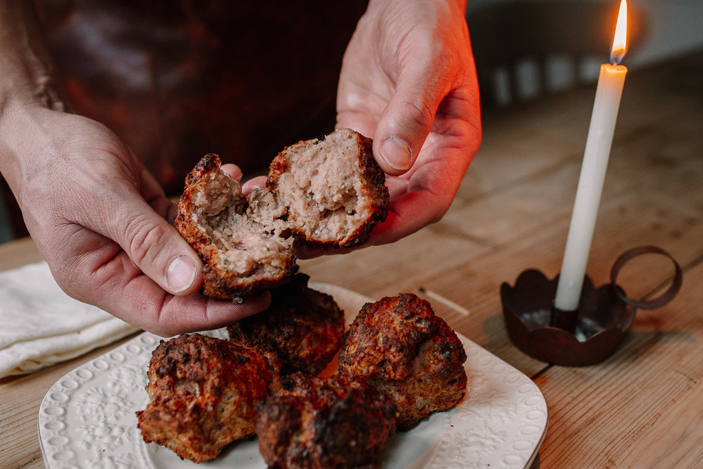 A close-up of a person's hands breaking open a cooked Pork Stuffing ball on a white plate, with other meatballs on the plate and a lit candle in a dark metal holder on a wooden table.