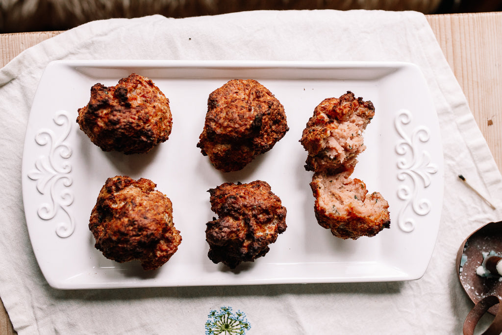 Six golden-brown cooked Pork Stuffing balls arranged on a white rectangular serving platter with decorative scrollwork, placed on a white cloth over a light wood surface.